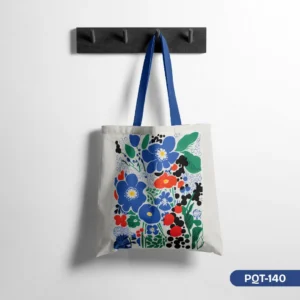 Scandi Pop Floral Tote Bag (PQT-140) showcasing a vibrant folk-art floral print in bold blues, reds, and greens on a light canvas with blue straps, hanging on a dark wood peg rail against a white wall.
