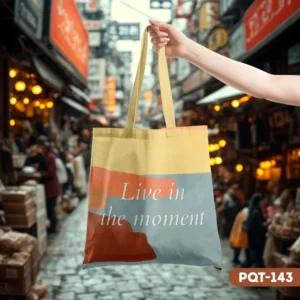 Whispering Path Tote Bag (PQT-143) with 'Live in the moment' quote, featuring earthy yellow, orange, and blue-grey abstract color blocks, held up by a hand in a blurred street market scene